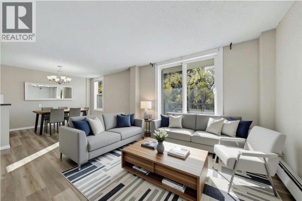 Living room with two grey sofas, coffee table, and view of trees through window. - 302, 1328 13 Avenue Sw, Calgary