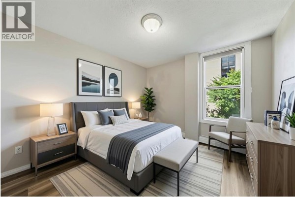 Bedroom with grey upholstered bed, nightstands, dresser, and framed artwork. - 302, 1328 13 Avenue Sw, Calgary