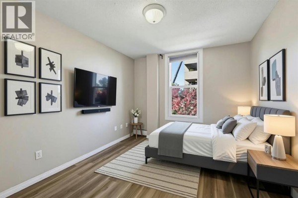 Bedroom with grey upholstered bed, wall-mounted TV, dresser, and window view. - 302, 1328 13 Avenue Sw, Calgary