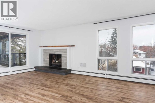 Living room with a gas fireplace, large windows overlooking trees, and light wood laminate flooring. - 302, 2611 15a Street Sw, Calgary