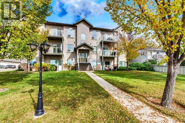 Side view of a beige apartment building entrance with a paved walkway through a grassy lawn. - 302, 7 Crystal Ridge Cove, Strathmore