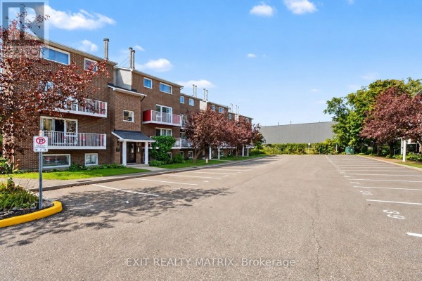 Exterior of brick apartment building with parking lot, trees, and distant industrial building. - 302 - 1589 St. Bernard Street, Ottawa
