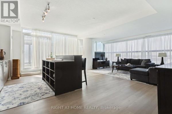 Spacious living and dining area with light wood floors, sheer curtains, and a dark kitchen island. - 302 - 3985 Grand Park Drive, Mississauga
