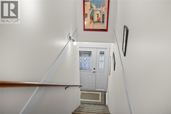 View down a staircase with a wooden handrail towards a white front door. - 302 1244 4th Ave, Ladysmith