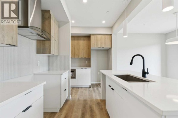 Contemporary kitchen with white countertops, light wood cabinets, stainless steel hood fan, and a central island with sink. - 302 Hotchkiss Common Se, Calgary
