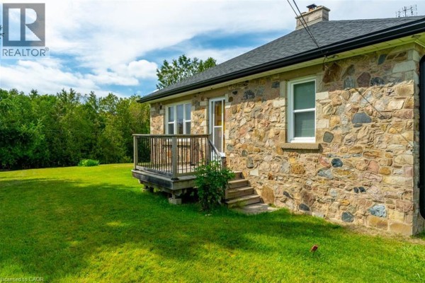 Side view of a stone house with a small wooden deck, steps, and windows, surrounded by green lawn. - 3029 Burnhamthorpe Road W, Milton