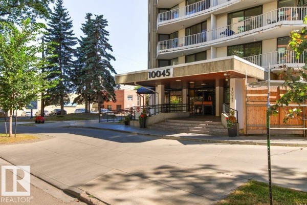 A wide exterior shot of a concrete apartment building surrounded by trees and parked cars. - #303 10045 117 St Nw, Edmonton
