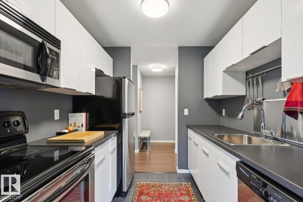 View down a narrow kitchen with black stovetop, stainless steel appliances, and white cabinets. - #303 10838 108 St Nw, Edmonton