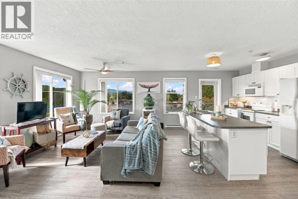 Living room and open-concept kitchen with gray sofa, patterned chairs, and white cabinets. - 303 129 Back Rd, Courtenay