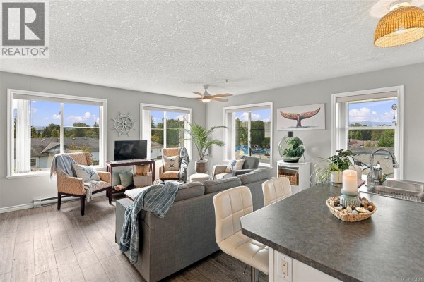 Living room and kitchen with gray sofa, island with bar stools, and white cabinets. - 303 129 Back Rd, Courtenay