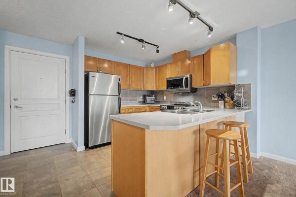 Apartment kitchen with light wood cabinets, stainless steel appliances, and island with stools. - #303 530 Hooke Rd Nw, Edmonton