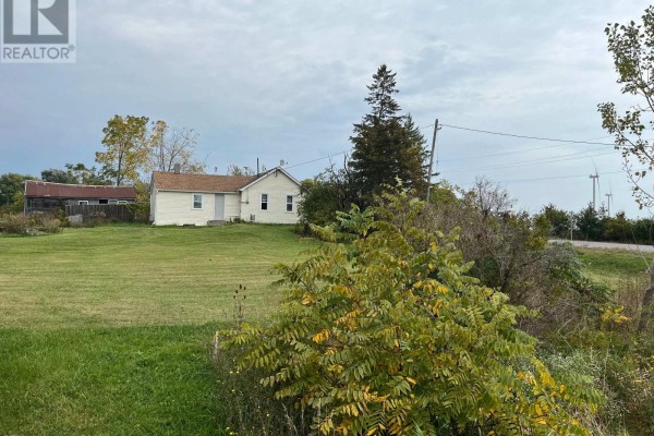 View of a white house from across a grassy field with trees and distant wind turbines. - 3031 Rainham Road, Haldimand