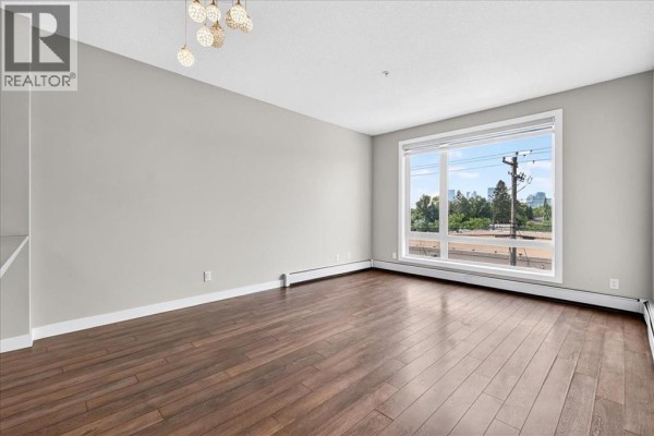 Living room with dark hardwood floors, light grey walls, and a large picture window overlooking the city. - 304, 607 17 Avenue Nw, Calgary