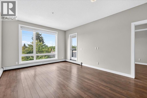 Spacious living area with dark hardwood floors, light grey walls, and a large window with tree views. - 304, 607 17 Avenue Nw, Calgary