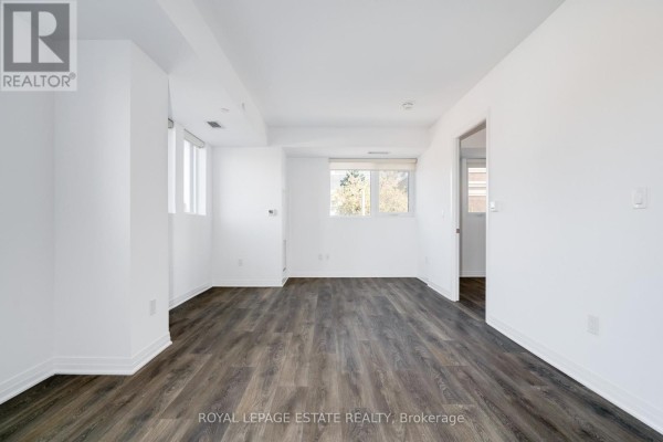 Empty living space with white walls, dark wood laminate flooring, and windows on two walls. - 304 - 2301 Danforth Avenue, Toronto