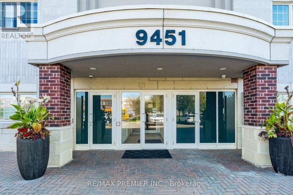Close-up of the glass entrance doors of apartment building 9451, flanked by brick pillars and potted plants. - 304 - 9451 Jane Street, Vaughan