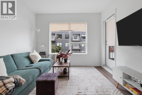 Living room with a teal sofa, wood coffee table, and a large window overlooking the building exterior. - 304 648 Lea Avenue, Coquitlam