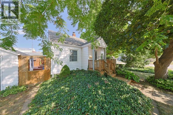 Side view of a white bungalow with a wooden deck and fence, partially obscured by trees. - 304 Lorne Avenue, Wallaceburg