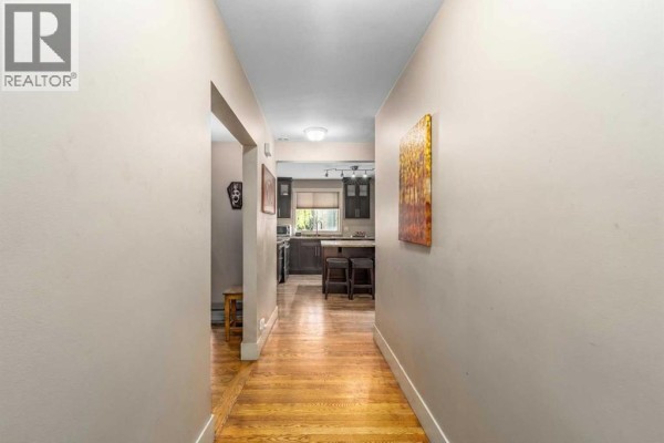 Hallway with hardwood floors, beige walls, and a view into a kitchen with dark cabinets. - 3042 28 Street Sw, Calgary