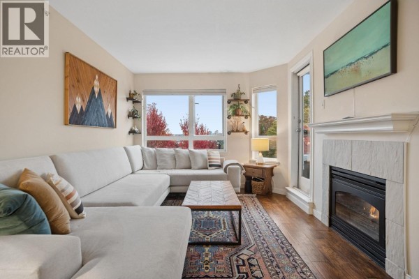 View of the living room sectional sofa, coffee table, rug, and fireplace with a window view. - 305 1707 Charles Street, Vancouver