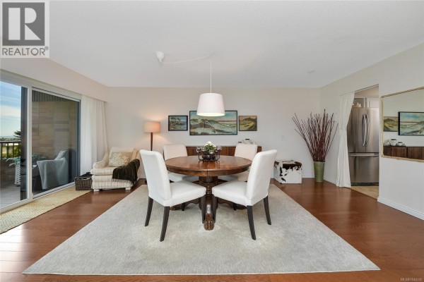 Dining room with a wooden table, white chairs, and a view of the kitchen. - 305 2930 Cook St, Victoria