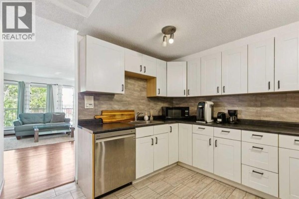 White kitchen with stainless steel appliances, black countertops, and a view of the living room. - 306, 1113 37 Street Sw, Calgary