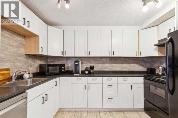 White kitchen with black appliances, dark countertops, stainless steel sink, and beige tiled floor. - 306, 1113 37 Street Sw, Calgary