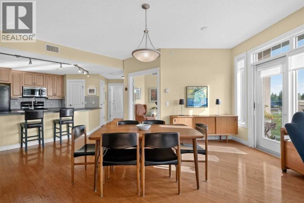 Dining area with wooden table, chairs, and view into a modern kitchen. - 306, 2121 98 Avenue Sw, Calgary
