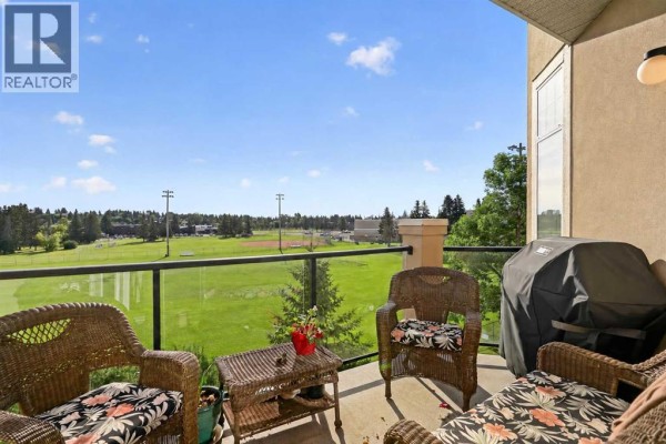 Covered balcony with wicker furniture, grill, and a view of a green field. - 306, 2121 98 Avenue Sw, Calgary