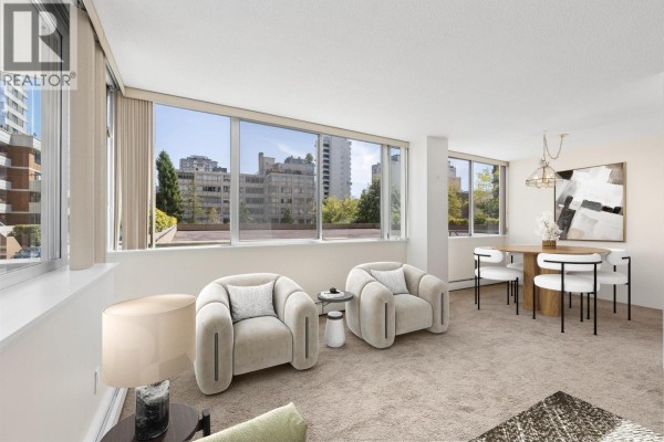 Living room with beige carpet, large windows, and view of buildings and trees. - 306 2055 Pendrell Street, Vancouver