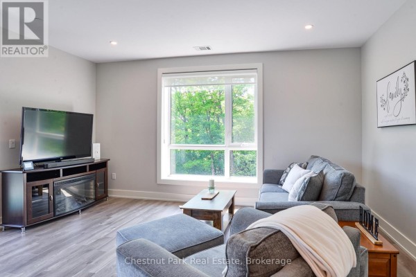 Living room with grey sofa, armchair, and large window overlooking trees. - 307 - 20 Campus Trail, Huntsville