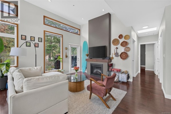 Living room with white sectional, gas fireplace, and large windows with forest views. - 307 170 Nursery Hill Dr, View Royal