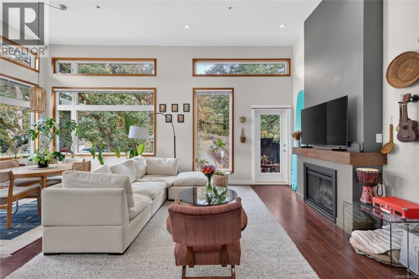 Living room with vaulted ceilings, large windows, fireplace, and a white sectional sofa. - 307 170 Nursery Hill Dr, View Royal
