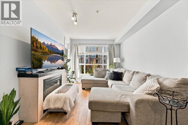 Living room with a large sectional sofa, electric fireplace, and hardwood floors, facing a large window. - 307 2343 Atkins Avenue, Port Coquitlam