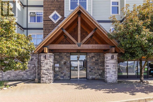 Close-up of the apartment building's wood- Beam entrance with stone pillars and glass doors, paved walkway in front. - 307 2881 Peatt Rd, Langford