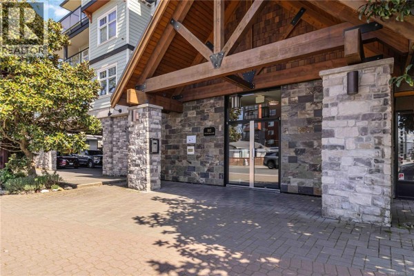 Close-up of the apartment building's wood-beam entrance with stone pillars and glass doors, shaded brick paving in front. - 307 2881 Peatt Rd, Langford