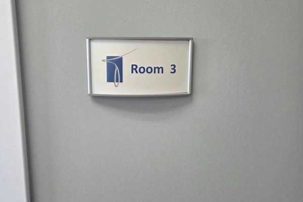 A rectangular sign on a grey wall reads "Room 3" with a blue logo. - 3075 Hospital Gate, Oakville