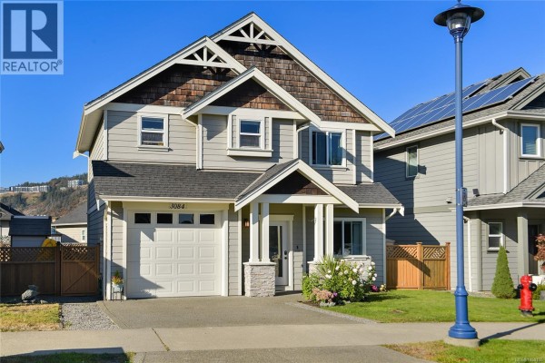 Close-up front view of a two-story house with beige siding, a white garage door, and decorative wood trim under the roof. - 3084 Langford Lake Rd, Langford