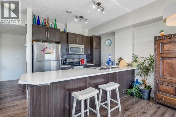Open concept kitchen and living area with island, dark brown cabinets, stainless steel appliances, and wood flooring. - 309, 250 Fireside View, Cochrane