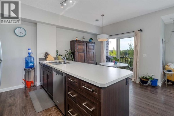 Kitchen island with sink and dishwasher, dark brown cabinets, and view of dining area and balcony door. - 309, 250 Fireside View, Cochrane