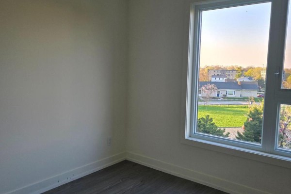 Empty room with white walls, dark wood laminate flooring, and a large window with residential views. - 309b - 16 Melbourne Avenue, St  Catharines