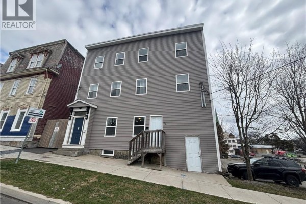 Front view of a grey multi-family building with wooden fence and parking area. - 31-33 Peters Street, Saint John