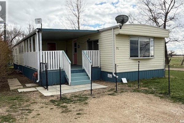 Exterior view of a cream-colored mobile home with a covered porch and satellite dish. Trees and grass surround it. - 31 Cypress Mobile Home Park, Maple Creek