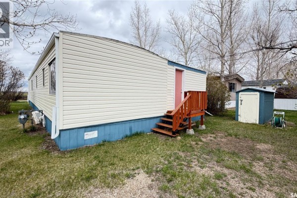 Rear right side of a mobile home with cream siding, blue skirting, and wooden steps leading to a pink door. - 31 Cypress Mobile Home Park, Maple Creek