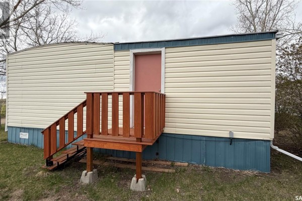 Back of the mobile home showing cream siding, blue skirting, and wooden steps with a railing. - 31 Cypress Mobile Home Park, Maple Creek