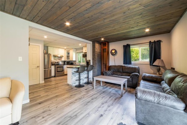 Living room and kitchen area with grey wood-look flooring, dark leather sofas, and a breakfast bar with stone accents. - 31 Gimli Road, Dunnottar