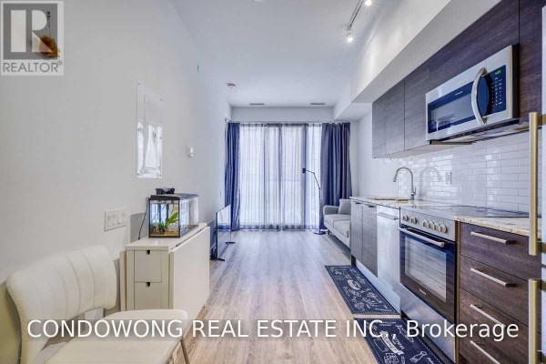 Living space with large windows, grey sofa, and open access to the kitchen area with wood cabinets. - 310 - 28 Wellesley Street E, Toronto