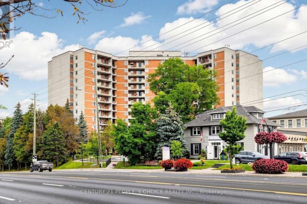 View of the brick apartment building from a street with trees and a smaller house nearby. - 310 - 7811 Yonge Street, Markham