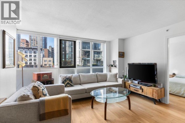 Living room with sectional sofa, glass coffee table, television, and view of city buildings from windows. - 310 1155 Seymour Street, Vancouver