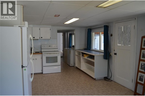 View of the kitchen and adjacent hallway, featuring white cabinetry and a window with blue curtains. - 310 Bancroft Street, Ashcroft